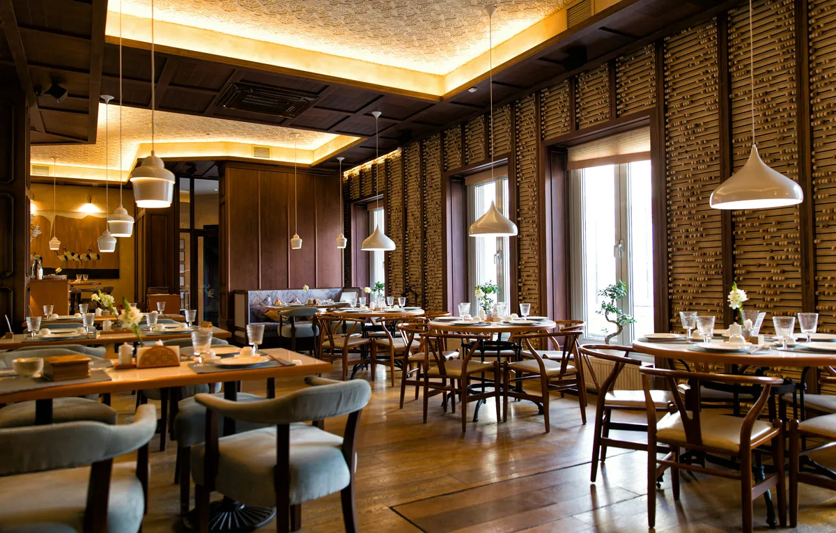 Salle de restaurant aux boiseries sombres, suspensions blanches, tables rondes et fauteuils en bois près de grandes fenêtres